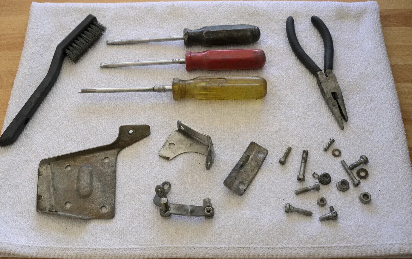 A wire brush, screwdrivers and slip-joint pliers laid out on a white towel beside hinge brackets, bolts and loose screws