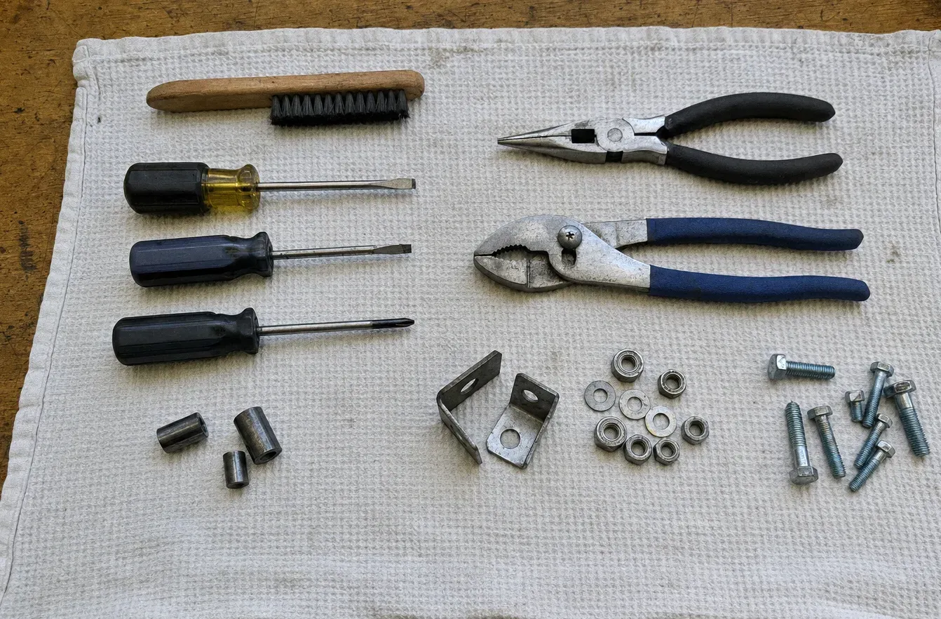 A wire brush, pliers and three screwdrivers on a white towel beside nuts, bolts and small metal brackets